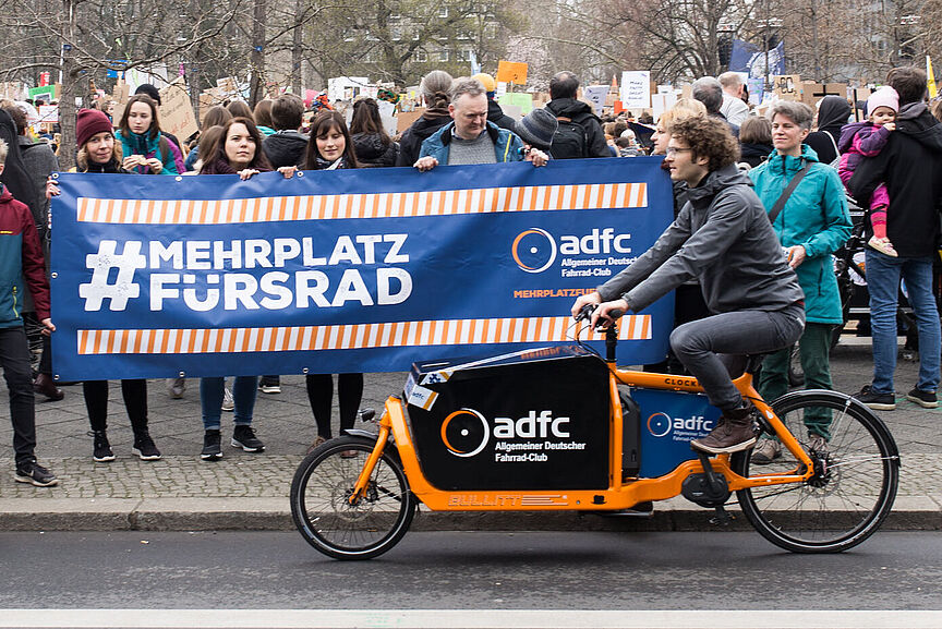 Ein Mensch auf einem Lastenrad vor einer Gruppe, die ein "Mehr Platz fürs Rad"-Banner hoch hält.