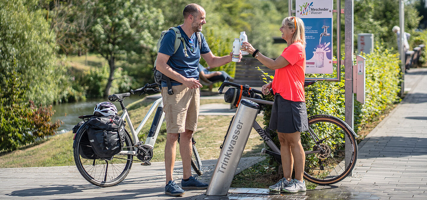 Wasserflaschen auffüllen am RuhrtalRadweg. Zwei Radfahrende füllen Trinkflaschen an einem Trinkbrunnen des RuhrtalRadwegs auf.