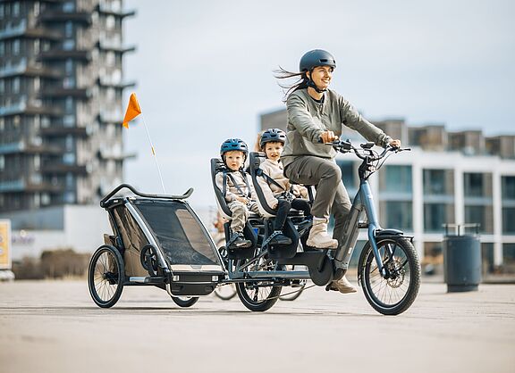 Frau fährt ein Lastenrad Longtail, auf dem langen Gepäckträger hinten sitzen zwei Kinder in je einem Kindersitz. Das Fahrrad zieht zudem einen Kinderfahrradanhänger.